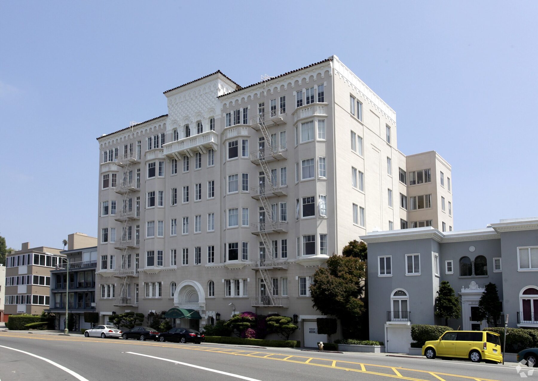 Six-story cream apartment building with bay windows, iron fire escapes and a tiled roof on a city boulevard